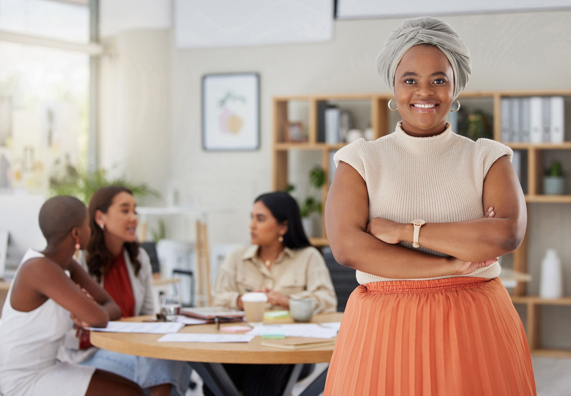 Portrait of smiling african american plus size business woman standing with her arms folded while colleagues sit behind her in office. Ambitious, confident, happy black professional with arms crossed